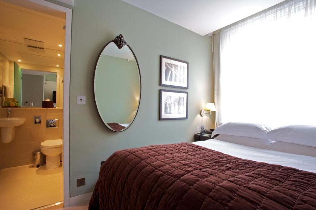 27 Best London Hotels with Balconies 2024 21 A room with a bed with red and white sheets beside an oval-shaped mirror near a bathroom. This is one of the rooms in the Ten Manchester Street Hotel.