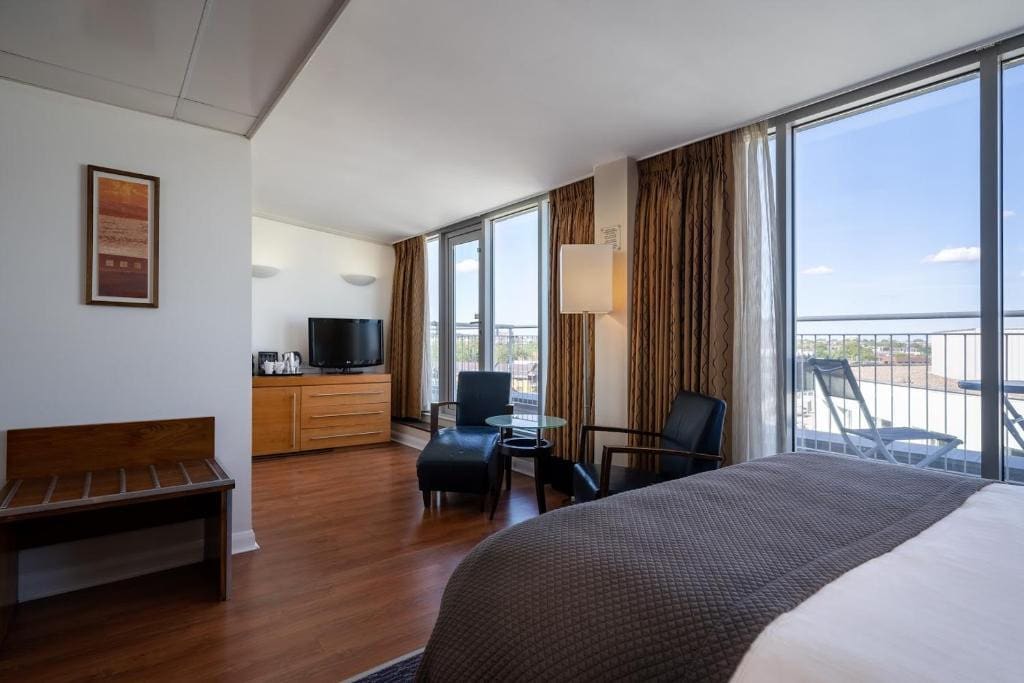 27 Best London Hotels with Balconies 2024 22 A hotel room with a large bed, in front of it is a round table with two upholstered chairs and a dresser with a TV on top. Beside the bed is a balcony with white railings and outdoor table and chairs. This is in the Holiday Inn London Camden Lock.