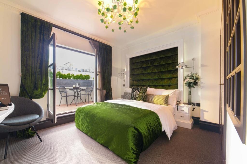 27 Best London Hotels with Balconies 2024 24 A bedroom with a green bed and a green headboard Beside it is a glass door to a balcony with metal furniture and a view of the city. This is in the Exhibitionist Hotel.