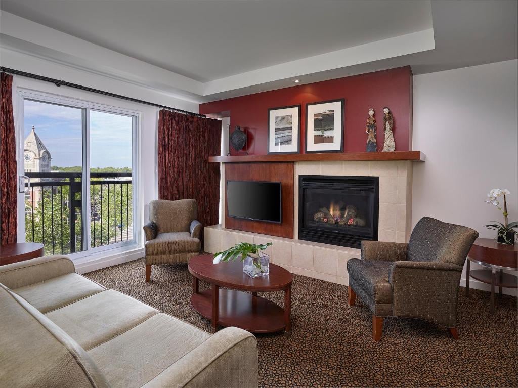 11 Best Edmonton Hotels With Balconies 2024 4 A white couch facing a wall-mounted flat screen TV, a fireplace, and two single upholstered chairs inside a carpeted room. A large balcony can be seen beside the couch. This one of the rooms in Metterra Hotel on Whyte