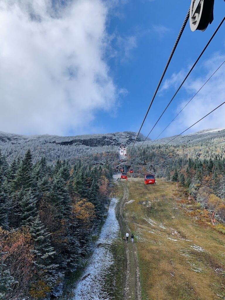 Why This World Traveler Chose to Live in Vermont Over All Other Places (Including Alaska!) 3 The view seen while enjoying the Gondola Skyride in Stowe, Vermont. The red gondola cars contrast against the fresh light dusting of snow and the golden late fall colors amongst the dark evergreen trees. The top of Mt. Mansfield can be seen in the distance. A couple with a toddler walk below on a grassy ski run.