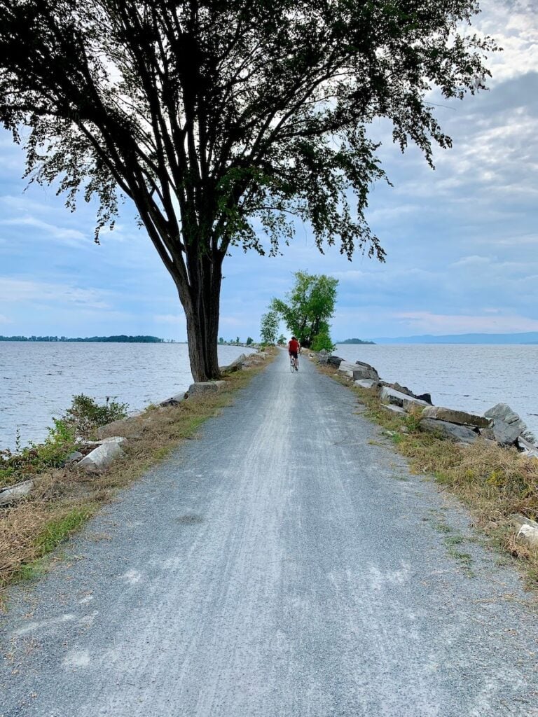 Why This World Traveler Chose to Live in Vermont Over All Other Places (Including Alaska!) 6 Cycling on the Causeway Path in Colchester, Vermont on a cloudy but bright day. The trail is edged by rocks and surrounded by water, Lake Champlain, on both sides. A lone deciduous tree stands in the foreground and a cyclist in a red shirt rides along under it.