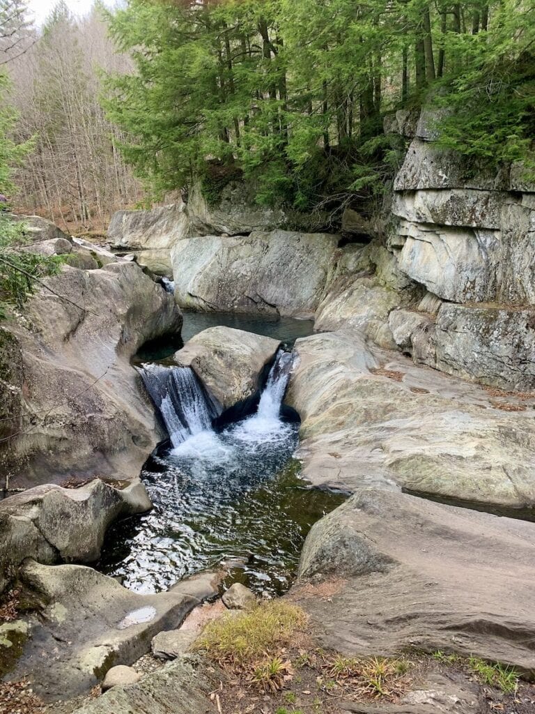 Why This World Traveler Chose to Live in Vermont Over All Other Places (Including Alaska!) 4 The view while standing on the bank of Warren Falls in Warren, Vermont. It is early November and all the leaves are off the trees. Dark evergreens tower above the pristine pools and the small waterfalls below in the Mad River. The granite boulders are smooth and weathered from running water.