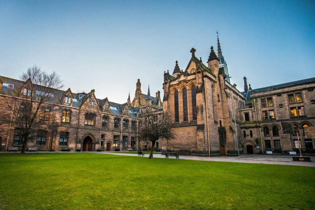 25 Pros and Cons of Living in Boston in 2024 1 Old architecture of Harvard. It's a green field in front of an orange brick building.