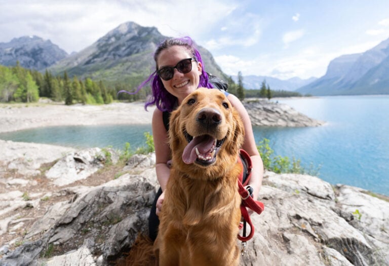 45 Pros and Cons of Living in Canada from a Canadian 1 Nina and Theo in Banff sitting beside a glacial lake on a hike. Theo the golden retriever sits in the foreground with his tongue out and Nina smiles over his shoulder with her sunglasses on and purple hair blowing in the wind