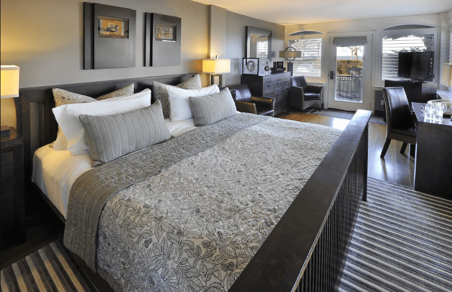 15 Luxurious Niagara Falls Resorts (2024 Guide) 6 Luxury interior of a spacious bedroom at Oban Inn with king bed and gray covers with 6 soft pillows and television. There is a balcony with view of the nature outside.