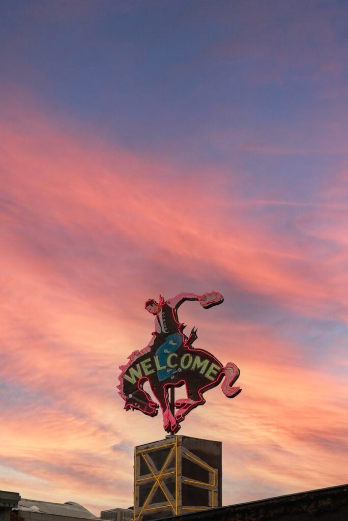 23 Pros and Cons of Living in Wyoming (2024) 1 red and black billboard of a cowboy on a bucking bronco in front of a pink sunset sky