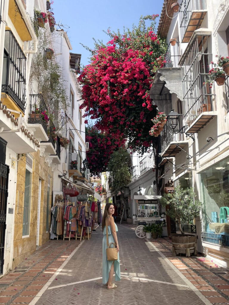 Discover Life Beyond the Tourist Trails in Spain's Coastal Towns Through a Local's Eyes 6 Woman walking through a charming street in Costa Del Sol, adorned with vibrant bougainvillea and local shops.