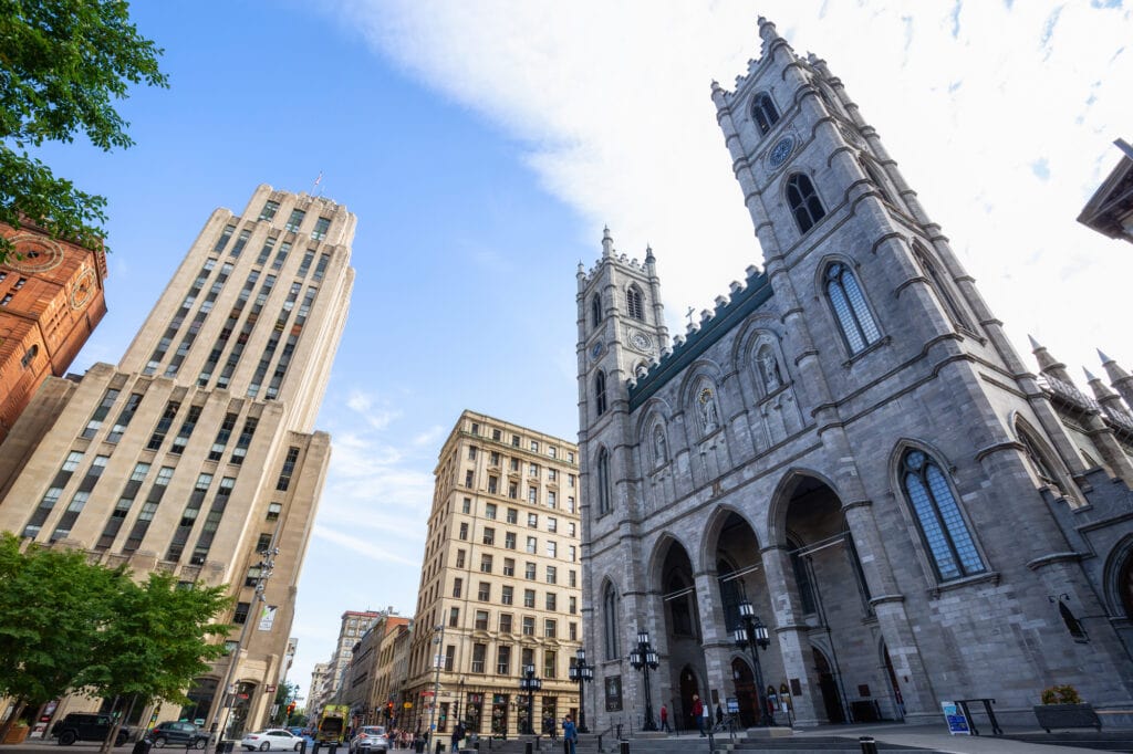 Notre Dame Quebec: Ultimate Visitor’s Travel Guide 2024 3 grey building of notre dame quebec with 2 towers and arched windows. the building is near 2 tall yellow buildings
