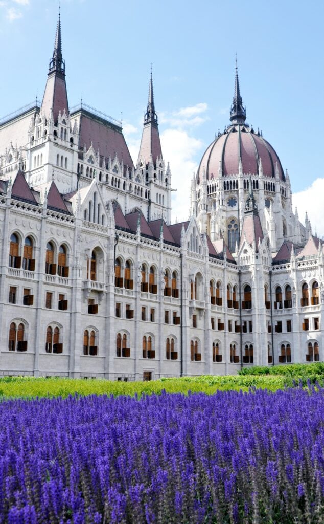 ALL European Countries With Digital Nomad Visas (2024) 10 The building of the Budapest Parliament in Hungary. The garden outside is surrounded with green and purple flowers.