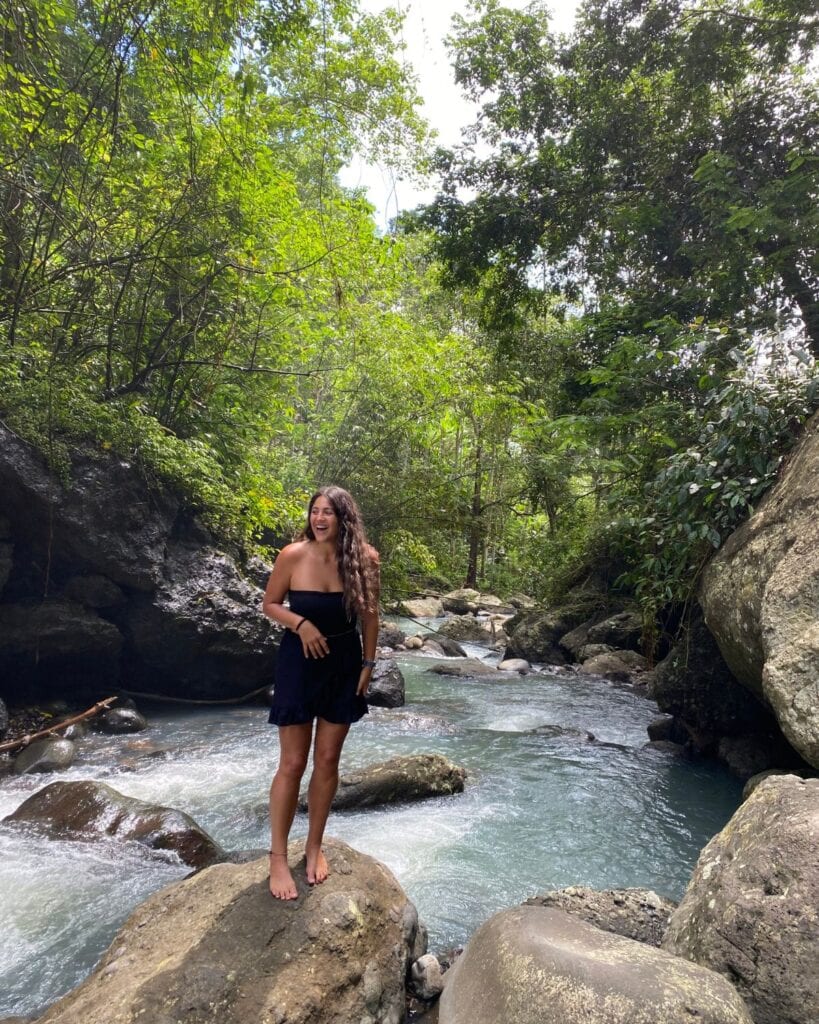 This Expat Traded London Life for Bali's Laidback Tropics 1 A smiling woman, Abi, in a black off-shoulder dress standing on a large rock in the middle of a gentle stream, surrounded by lush greenery and large boulders in a tropical forest setting in Bali.