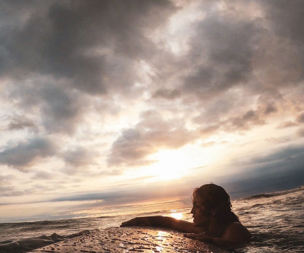 This Expat Traded London Life for Bali's Laidback Tropics 3 Abi holding onto a surfboard as she floats in the water silhouetted at golden hour in Bali.