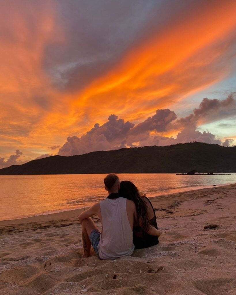 This Expat Traded London Life for Bali's Laidback Tropics 2 Abi and her partner sitting on a sandy beach in Bali looking at the orange sunset over the water with his arm around her.
