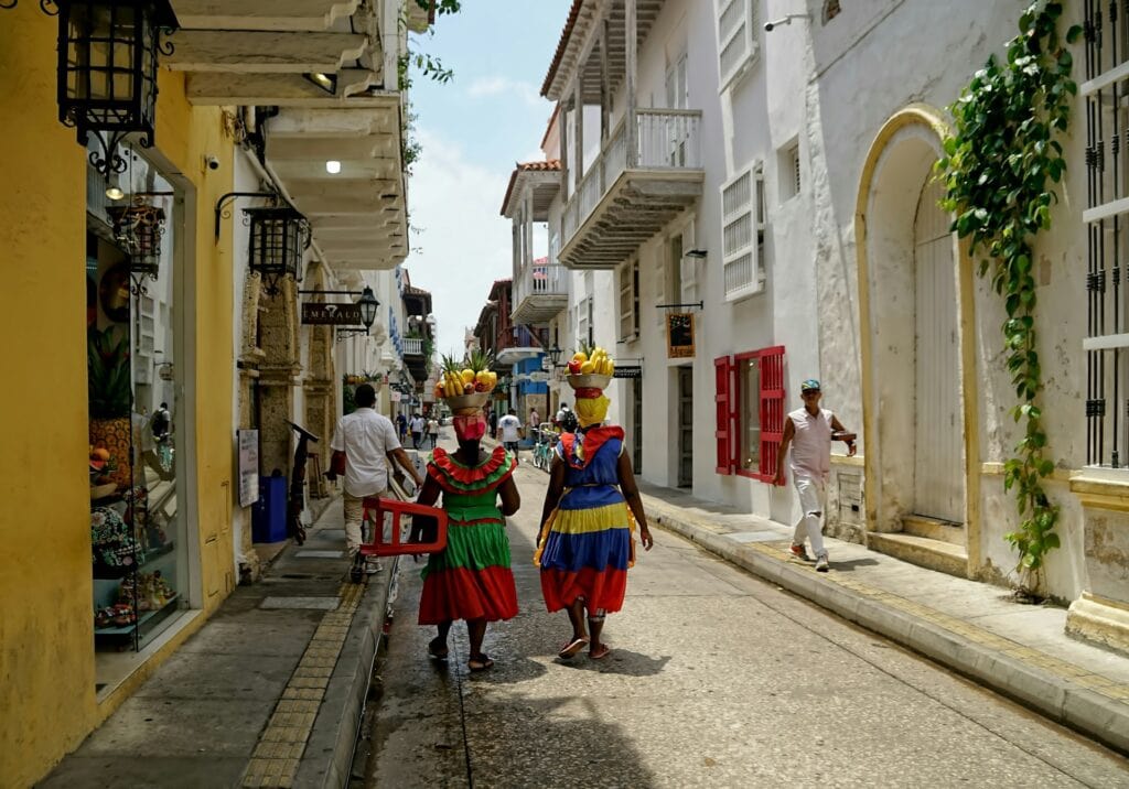 Top 17 Countries With Retirement Visas (2024 Guidelines) 6 Two woman wearing colorful dresses walking along the street while carrying fruits in a basket above their heads.
