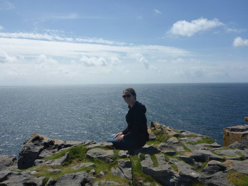 13 Countries That Will Pay You To Move There (Expat’s Picks) 7 A girl wearing sunglasses sitting on the edge of a cliff overlooking the seea in an island in Ireland.