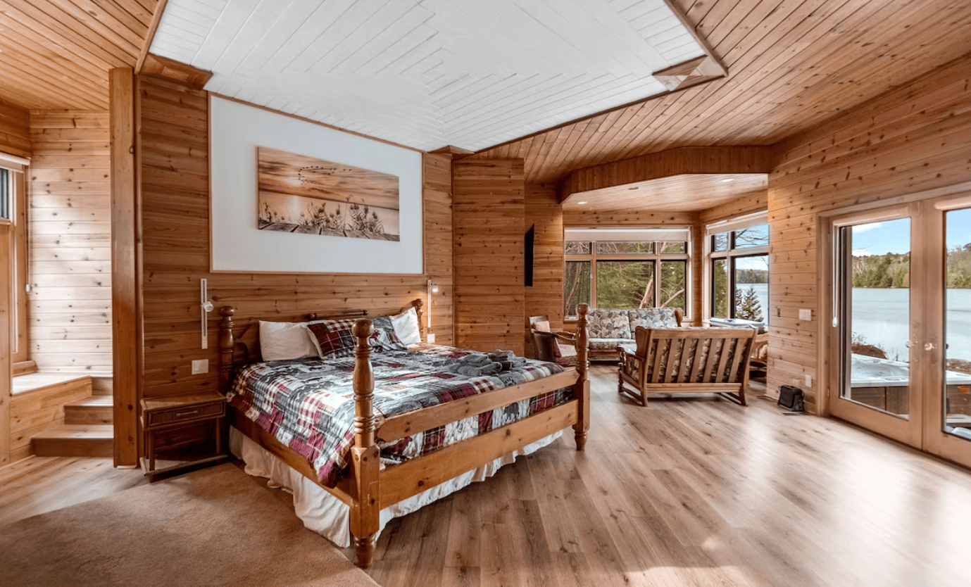 20 Top Airbnb Cottages in Muskoka for an Epic Getaway 59 the all wooden cabin interior, including a bed with large windows and a sitting area looking out towards the lake