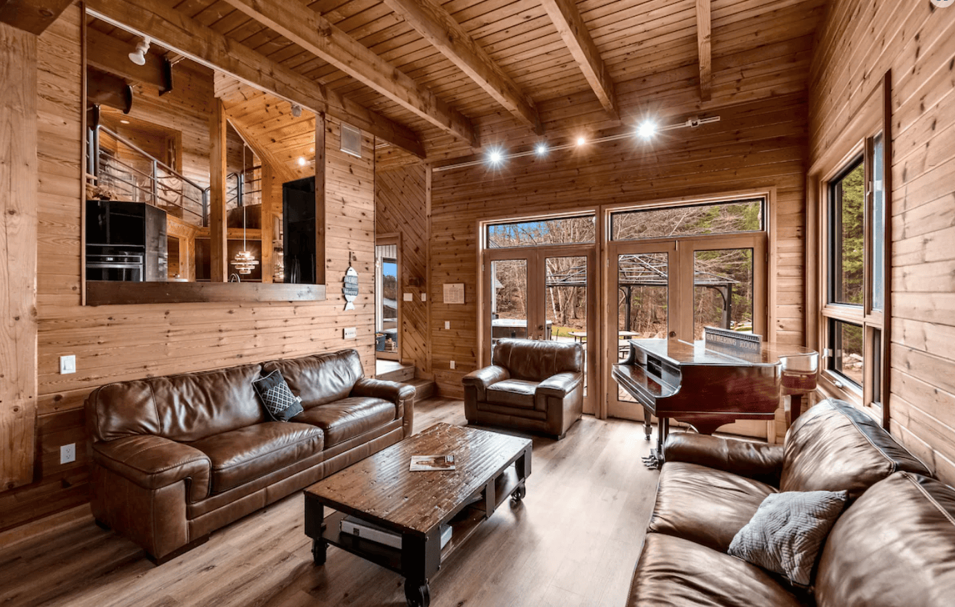 20 Top Airbnb Cottages in Muskoka for an Epic Getaway 60 a living space with brown leather couches, a piano, and great natural lighting from the large windows.