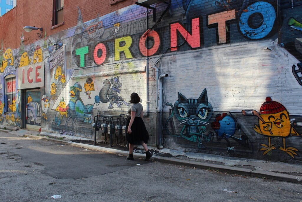 Best Time to Visit Toronto (A Month-By-Month Guide) 1 Nina walking through Graffiti Alley with a variety of small street art paintings on the brick wall.