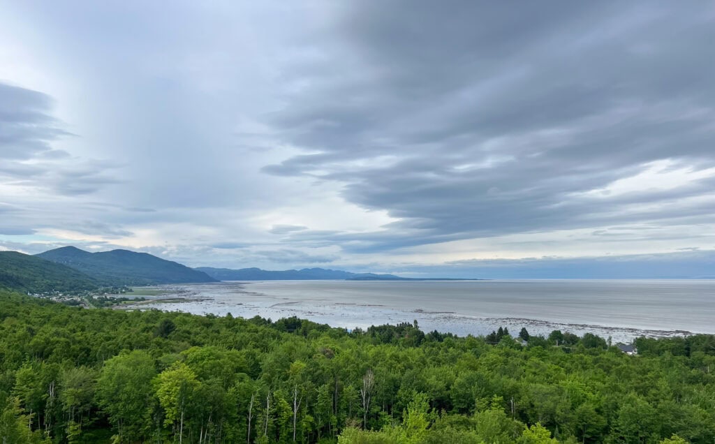 My 4 Day Charlevoix Itinerary to Recharge in Quebec 4 Wide view of the St. Lawrence River with rolling green hills and scattered clouds under a moody sky near Club Med Charlevoix. A scenic lookout point on the drive through the region.