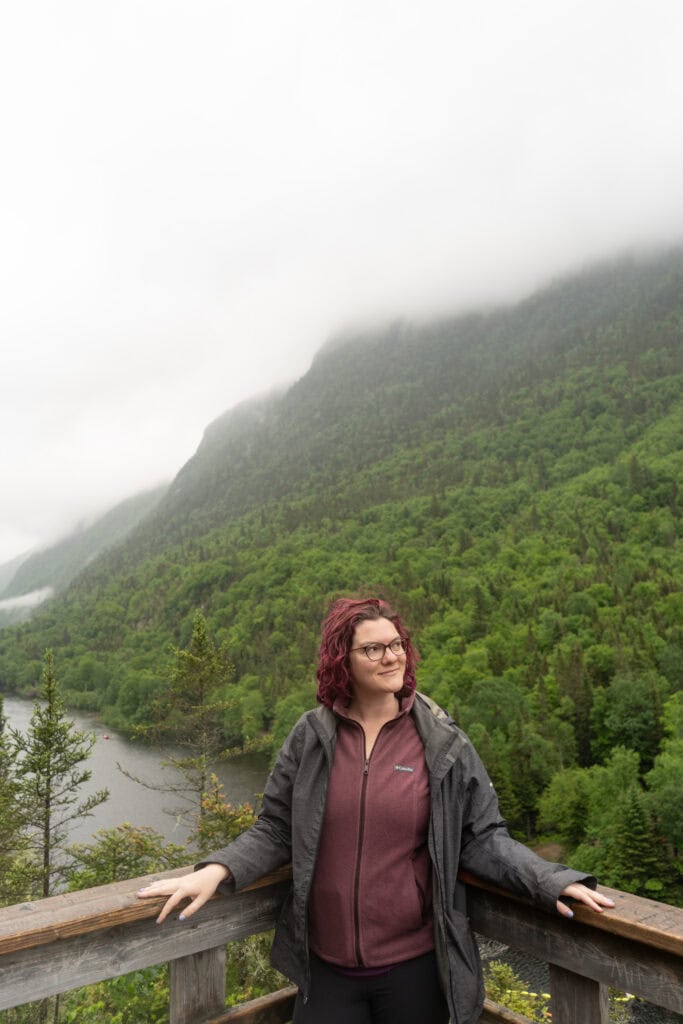 My 4 Day Charlevoix Itinerary to Recharge in Quebec 11 Nina with short purple hair and glasses standing at a wooden lookout railing with misty green mountains behind them in Hautes-Gorges-de-la-Rivière-Malbaie National Park. A peaceful stop on a Charlevoix itinerary for nature lovers.