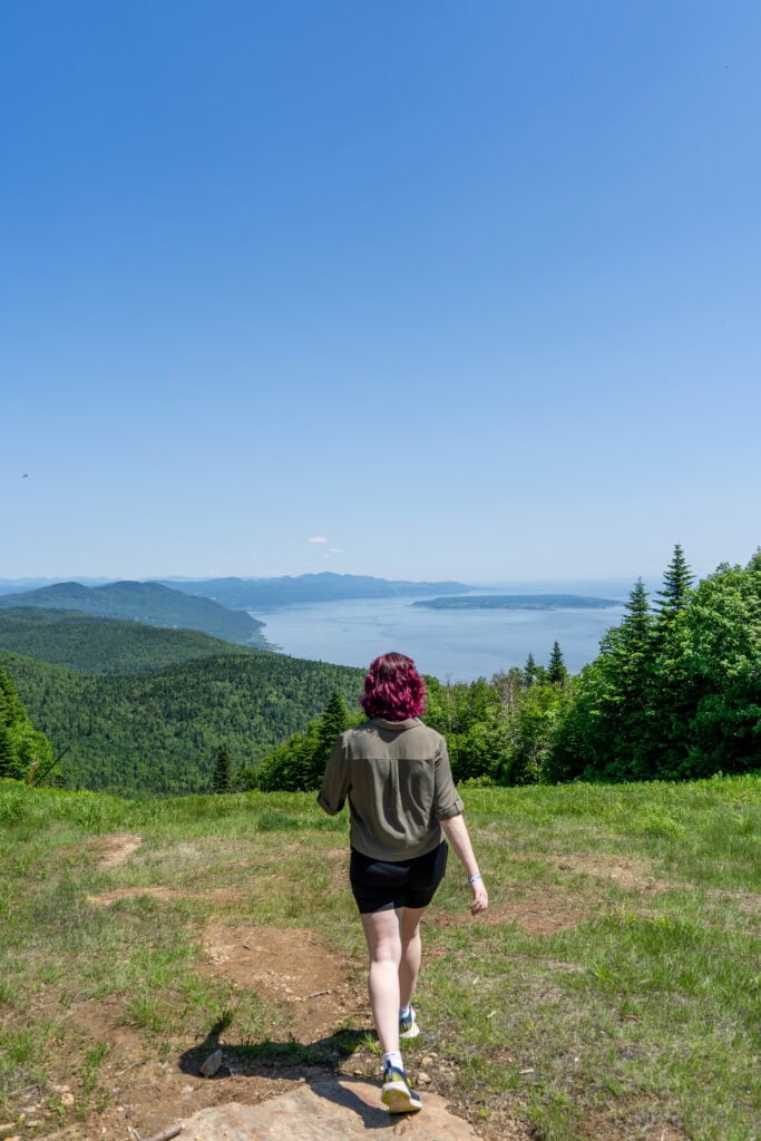 My 4 Day Charlevoix Itinerary to Recharge in Quebec 6 Nina walking away on a grassy trail overlooking lush green hills and the St. Lawrence River at Le Massif de Charlevoix.